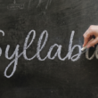 A woman holds a piece of chalk and finishes writing the word "Syllabus" in cursive across a dark, dusty chalkboard.