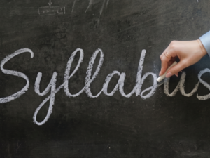 A woman holds a piece of chalk and finishes writing the word "Syllabus" in cursive across a dark, dusty chalkboard.