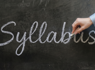 A woman holds a piece of chalk and finishes writing the word "Syllabus" in cursive across a dark, dusty chalkboard.