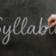 A woman holds a piece of chalk and finishes writing the word "Syllabus" in cursive across a dark, dusty chalkboard.